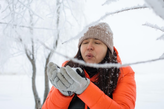 Woman of middle age behind some twigs covered with snow blowing snow off gloves on walk in snowy forest with tree in background Magic winter time full of white color