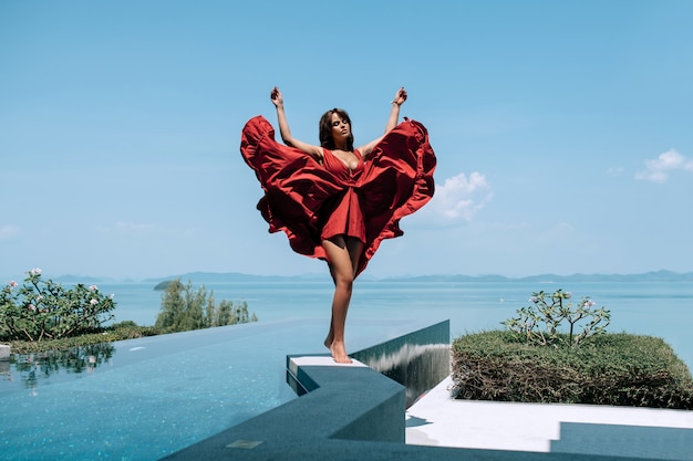 Woman model in fashion red dress standing  on the  edge of infinity swimming pool with sea view. 