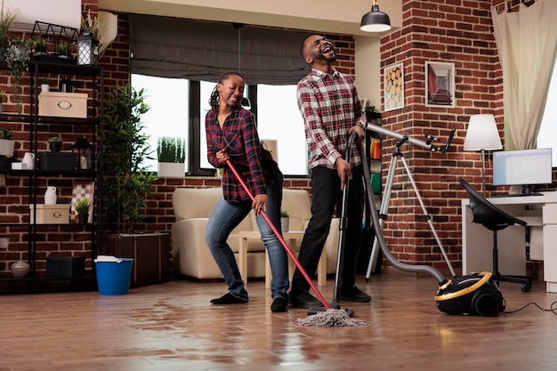 Woman mopping the floor and dancing while man sings with passion holding vacuum cleaner. African american couple cleaning house on weekend, taking care of keeping home tidy and clean.