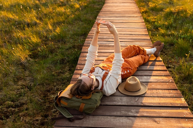 Photo woman naturalist orange overalls makes selfie on the phone resting lying on backpack on wooden path