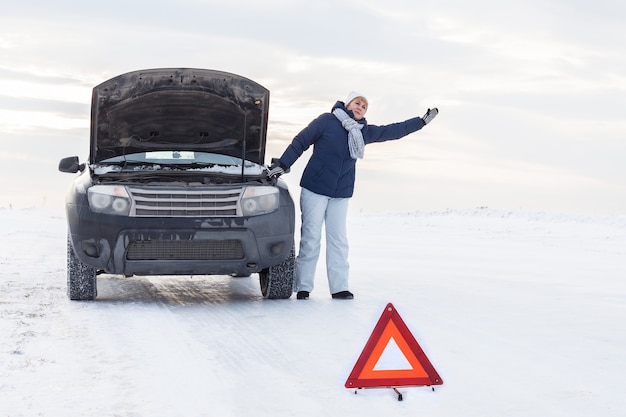 A woman near a broken car. She's asking for help. Around winter and snow field. There is an emergency sign.