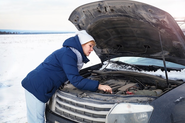 A woman near a broken car trying to fix the engine. She's cold. Around winter and snow field.