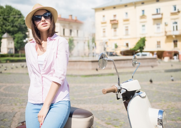 Woman in nifty hat standing near scooter outdoors.