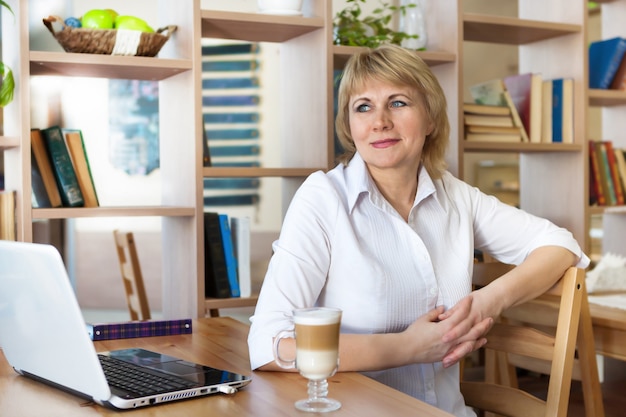 A woman in the office is working on a laptop. She smiles. A middle-aged woman, an adult in a cafe.