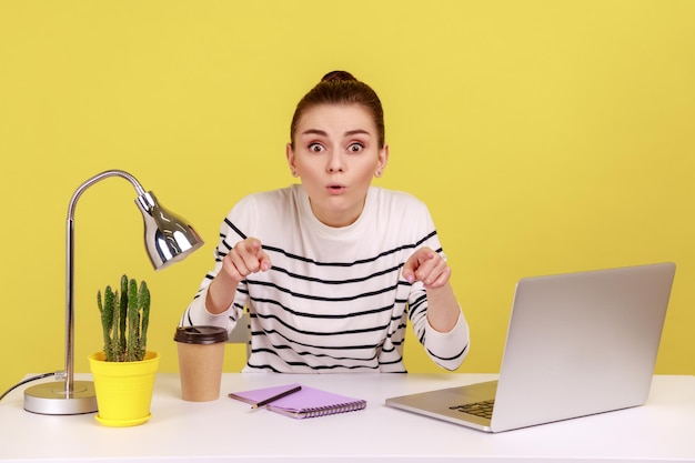 Woman office worker sitting workplace looking and pointing at camera with astonished surprised face