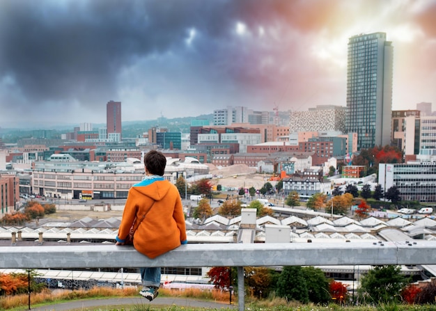 A woman in an orange coat sitting back to the camera in front of the city an autumn evening