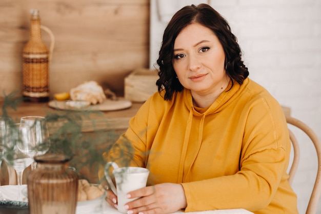 A woman in an orange hoodie is sitting at a table in the kitchen drinking coffee.