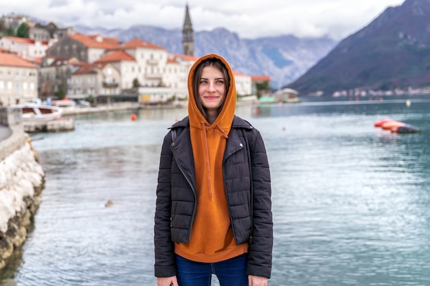 Woman in orange hoodie stands by the sea in city Perast on the background in Montenegro