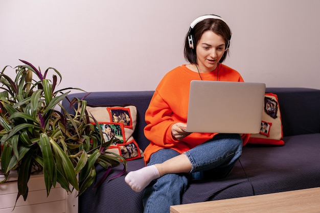 A woman in an orange jacket and blue jeans on the couch sits a woman at the computer in headphones