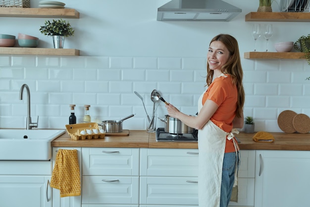 a woman in an orange shirt stands in a kitchen with a pot of food