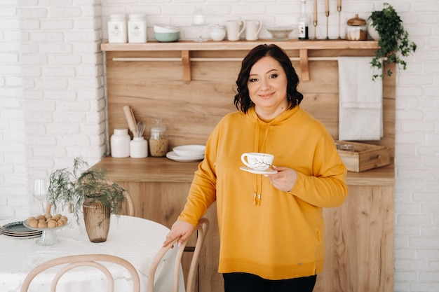 A woman in an orange sweatshirt drinks coffee in the kitchen at home.