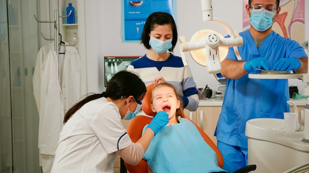 Woman orthodontist lighting the lamp examining little patient using sterile dental tools in stomatological clinic. Dentistry doctor speaking to kid sitting on stomatological chair, girl opening mouth