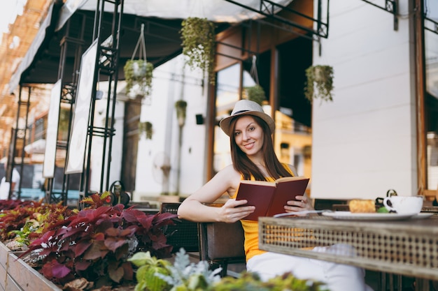 Woman in outdoors street coffee shop cafe sitting at table in hat, reading book with cup of cappuccino, cake, relaxing in restaurant during free time