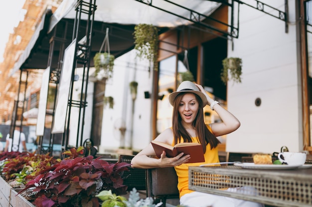 Woman in outdoors street coffee shop cafe sitting at table in hat, reading book with cup of cappuccino, cake, relaxing in restaurant during free time