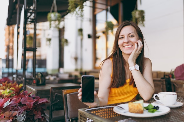 Woman in outdoors street coffee shop cafe sitting at table with cup of tea, cake, hold in hand mobile phone with blank empty screen