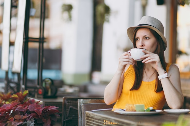 Woman in outdoors street coffee shop cafe sitting at table in yellow clothes hat with cup of cappuccino, cake, relaxing in restaurant during free time