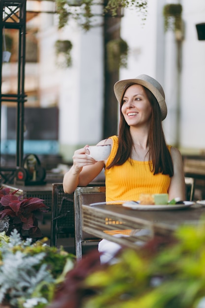 Woman in outdoors street coffee shop cafe sitting at table in yellow clothes hat with cup of cappuccino, cake, relaxing in restaurant during free time