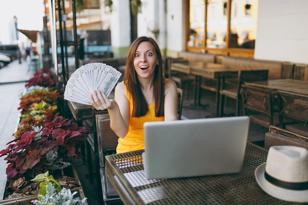 Woman in outdoors street coffee shop cafe sitting with modern laptop pc computer, holds in hand bunch of dollars banknotes