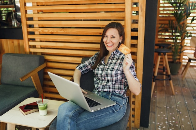 Woman in outdoors street summer coffee shop wooden cafe sitting working on laptop pc computer, hold bank credit card relaxing during free time