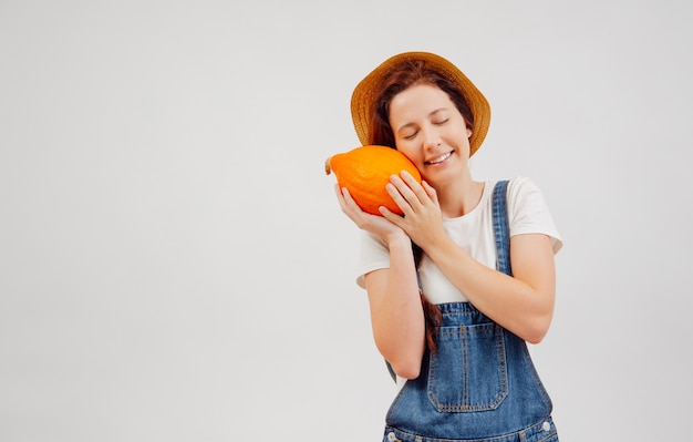 Woman in overalls and a hat presses a ripe orange pumpkin on a white background