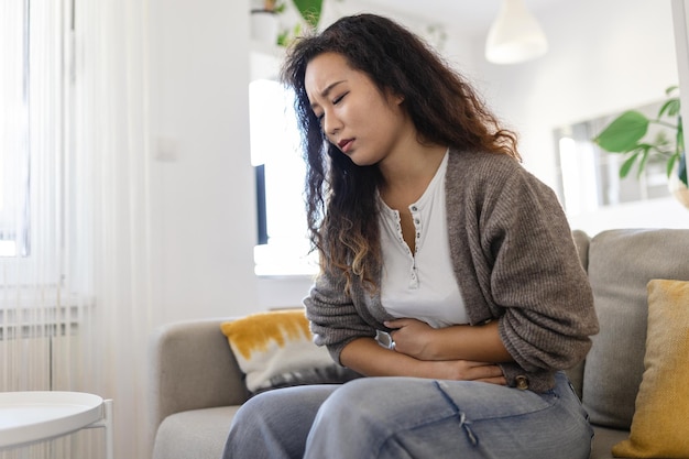 Woman in painful expression holding hands against belly suffering menstrual period pain lying sad on home bed having tummy cramp in female health concept