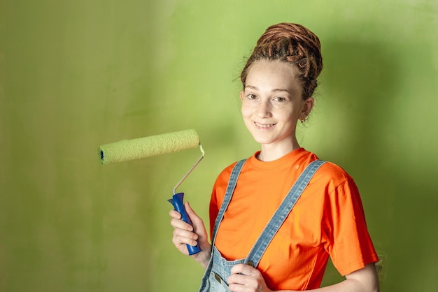 Woman painter with dreadlocks in an orange Tshirt and a denim jumpsuit make repairs in the apartment and paints the wall green with a roller