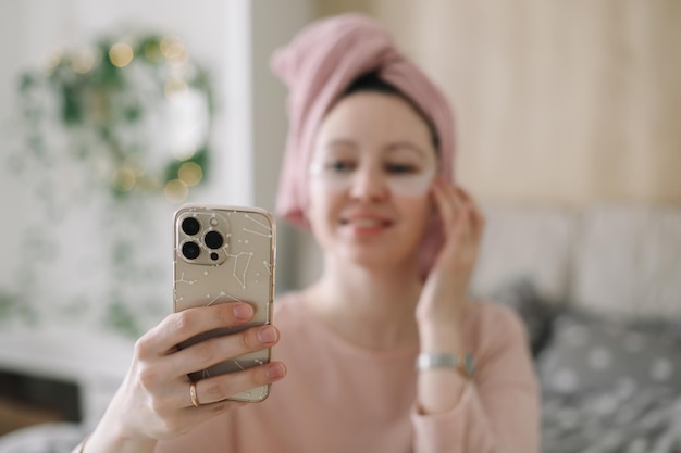 Woman in pajama with eyes patches and towel on head using phone in bed Morning habits and home beauty routine