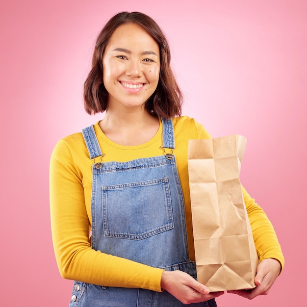 Woman paper bag and studio portrait for grocery shopping discount or choice with smile by pink background Japanese gen z girl retail customer and package for product sale or happy for cheap deal