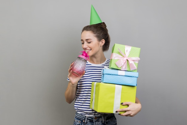 Woman in party cone holding stack of present boxes drinking cocktail enjoying birthday party