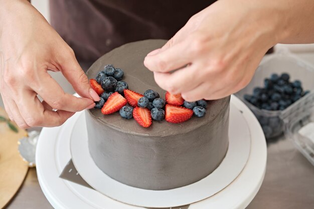 Woman pastry chef decorates grey cake with berries closeup Cake making process Selective focus