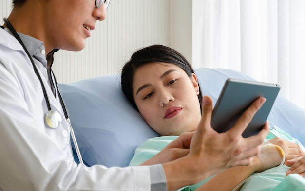 Woman patient on hospital bed looking at digital tablet while male doctor explain and discuss her health concerns