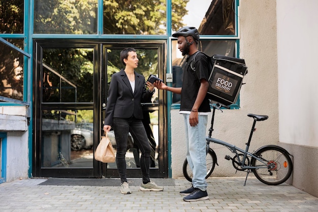 Woman paying for office food delivery service with smartphone, contactless pos terminal, mobile wallet payment. Courier delivering restaurant takeaway meal employee in front of company building