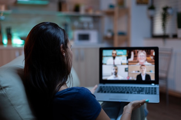 Woman in pijamas lying on sofa using laptop talking about sale report in video conference with team. Remote worker having online meeting consulting with colleagues using videocall and webcam chat