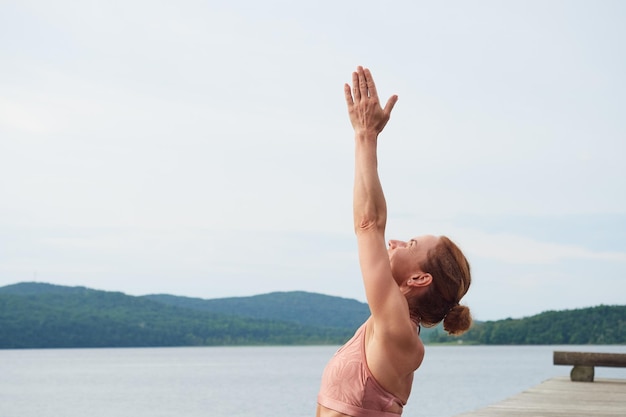 Woman in pink sportswear is training on the seashore raising her hands up