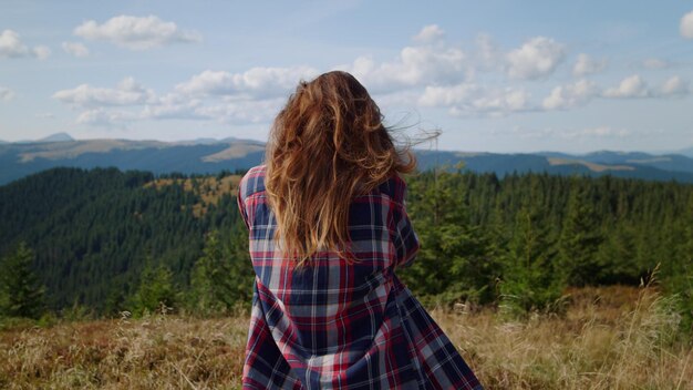 A woman in a plaid dress stands on a hill with mountains in the background.