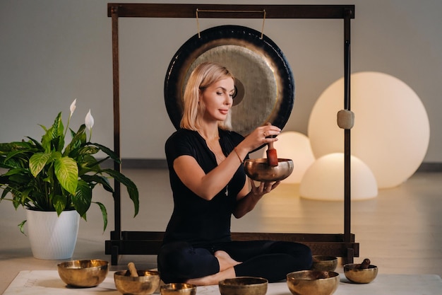 A woman plays a Tibetan singing bowl while sitting on a yoga mat against the background of a gong