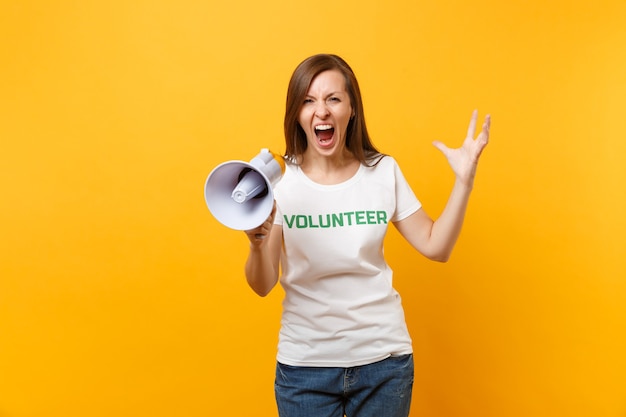 Woman portrait in white t-shirt written inscription green title volunteer scream in public address megaphone isolated on yellow background. Voluntary free assistance help, charity grace work concept.