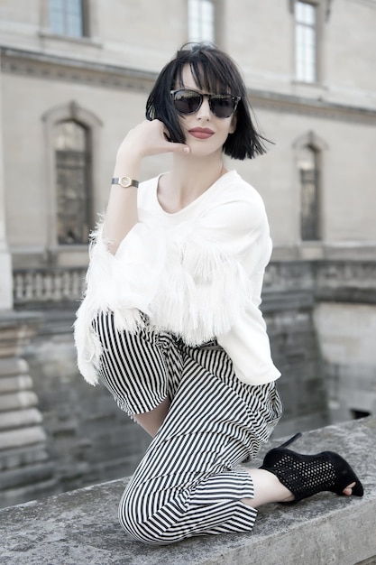 Woman pose on stone fence on house facade. Model in high heel shoes, fashionable clothes. Fashion, vogue concept. Girl with brunette hair, red lips in sunglasses. Beauty, look, style, parisian woman