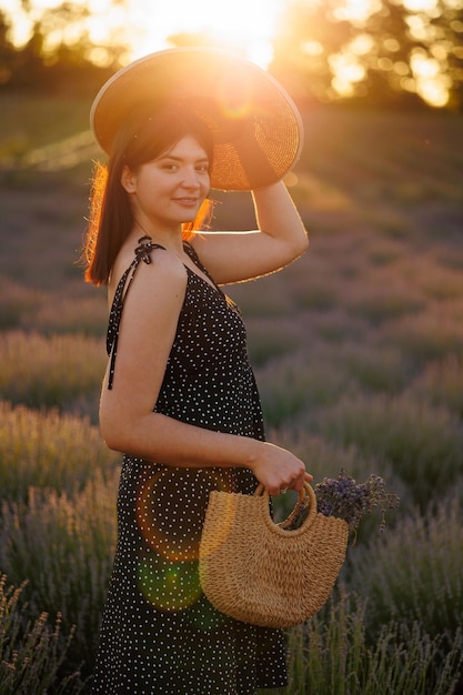 Woman posing in a lavender field with a straw hat and wicker bag at sunset