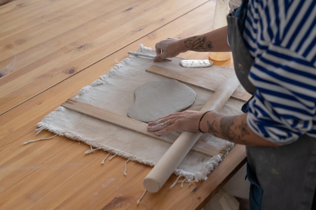 Woman potter artist in apron use wooden rolling pin to roll out clay making handmade utensils