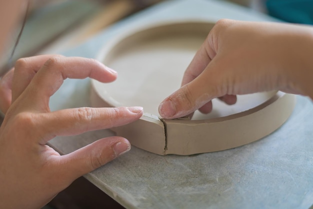 Woman potter working on potters wheel making ceramic pot from clay in pottery workshop art concept Focus hand young woman attaching clay product part to future ceramic product Pottery workshop