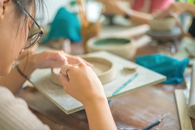 Woman potter working on potters wheel making ceramic pot from clay in pottery workshop art concept Focus hand young woman attaching clay product part to future ceramic product Pottery workshop