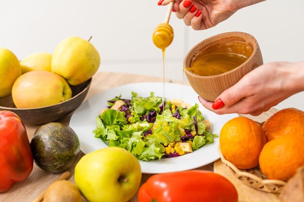 Woman pouring honey mustard dressing into bowl with fresh salad on table.