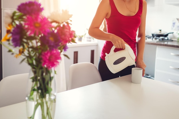 Woman pouring hot water from electric kettle in kitchen girl making tea modern kitchen design