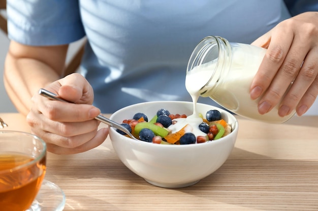 Woman pouring tasty yogurt onto fruits in bowl at table