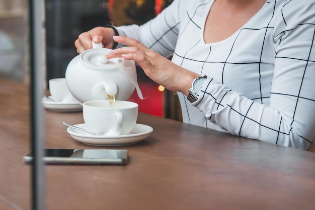 Woman pouring tea from white kettle to cap in cafe