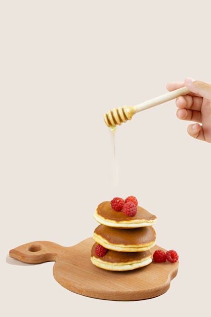 A woman pours honey on a stack of pancakes with red raspberries on the board