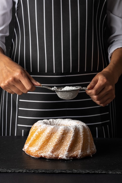 Woman powder of sugar to vanilla bundt cake on dark background.