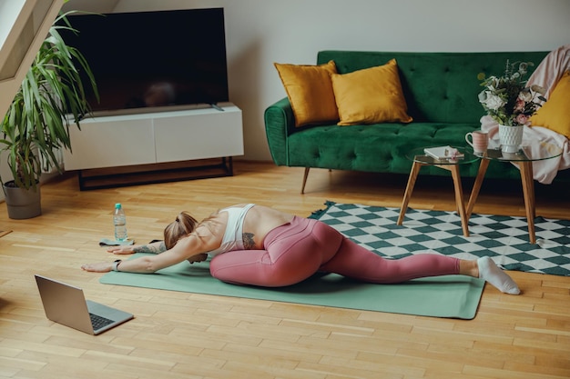 A woman practices yoga on hardwood flooring in her house with a laptop nearby