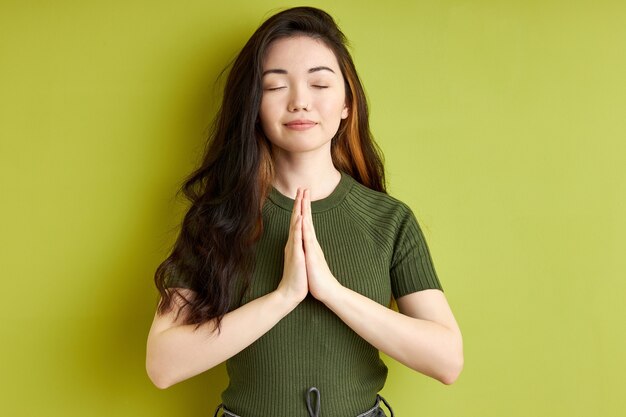 Woman praying with hands together asking for forgiveness smiling with eyes closed, pleasant brunette female isolated over green background